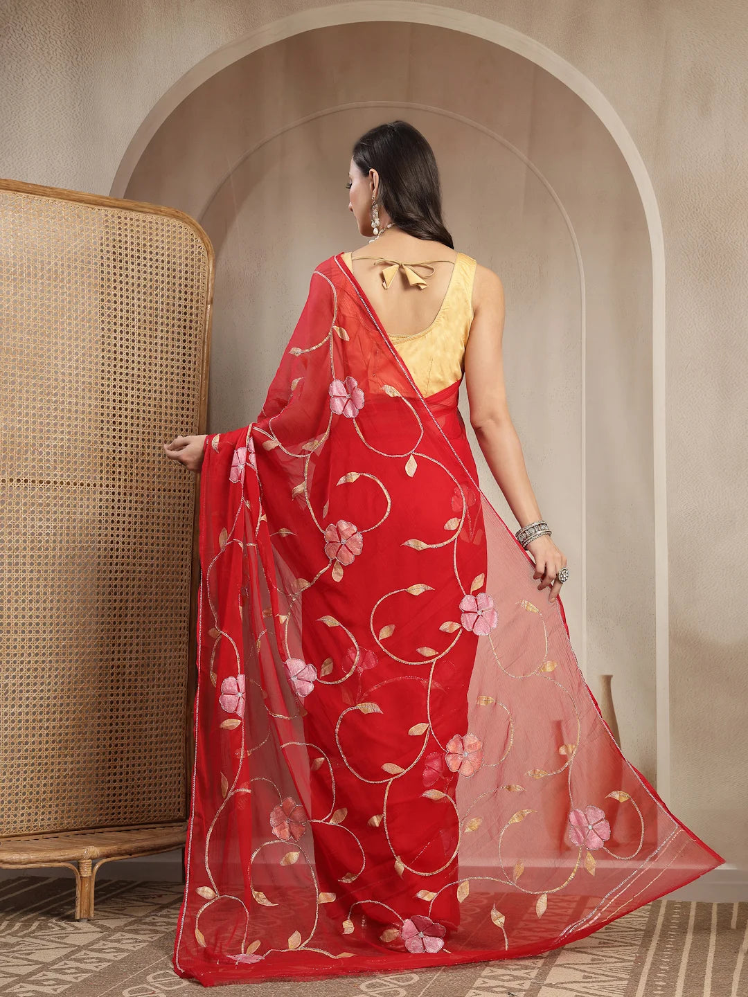 A woman wearing a red saree with pink floral embroidery and a yellow blouse, standing in front of a beige arched wall next to a woven cane screen.