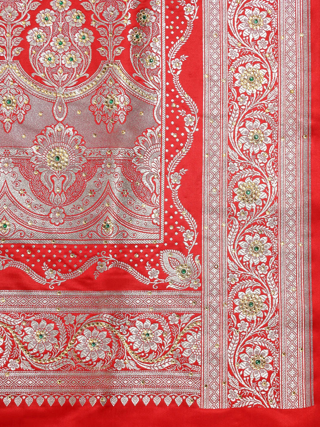 A close-up of a red and silver patterned fabric, likely a Banarasi silk saree or textile, featuring intricate floral and paisley motifs with small green and gold accents.