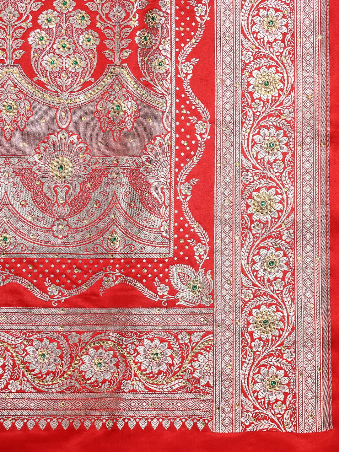 A close-up of a red and silver patterned fabric, likely a Banarasi silk saree or textile, featuring intricate floral and paisley motifs with small green and gold accents.