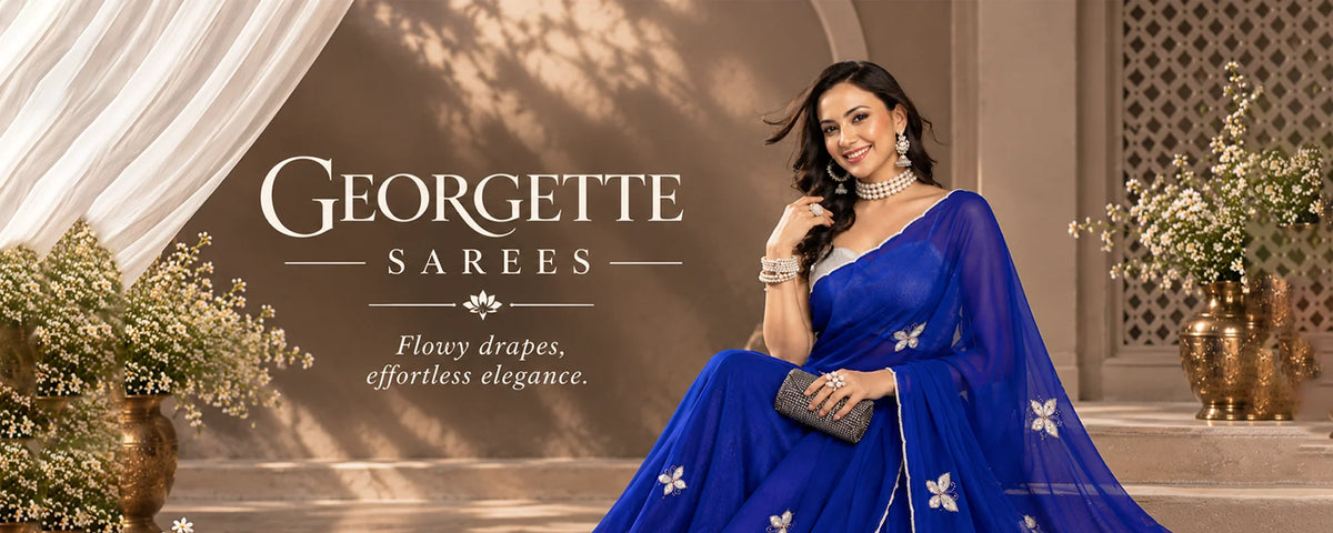 A woman models a royal blue sukunj Georgette saree with white floral embroidery, seated in an elegant indoor setting with draped curtains and floral arrangements. The brand name 'Georgette Sarees' is displayed on the left side of the image.