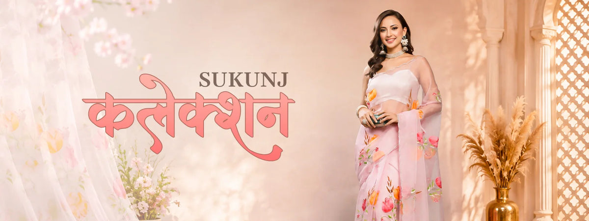 A woman modelling a light pink floral saree with a white blouse, standing in a warm, decorated indoor setting.